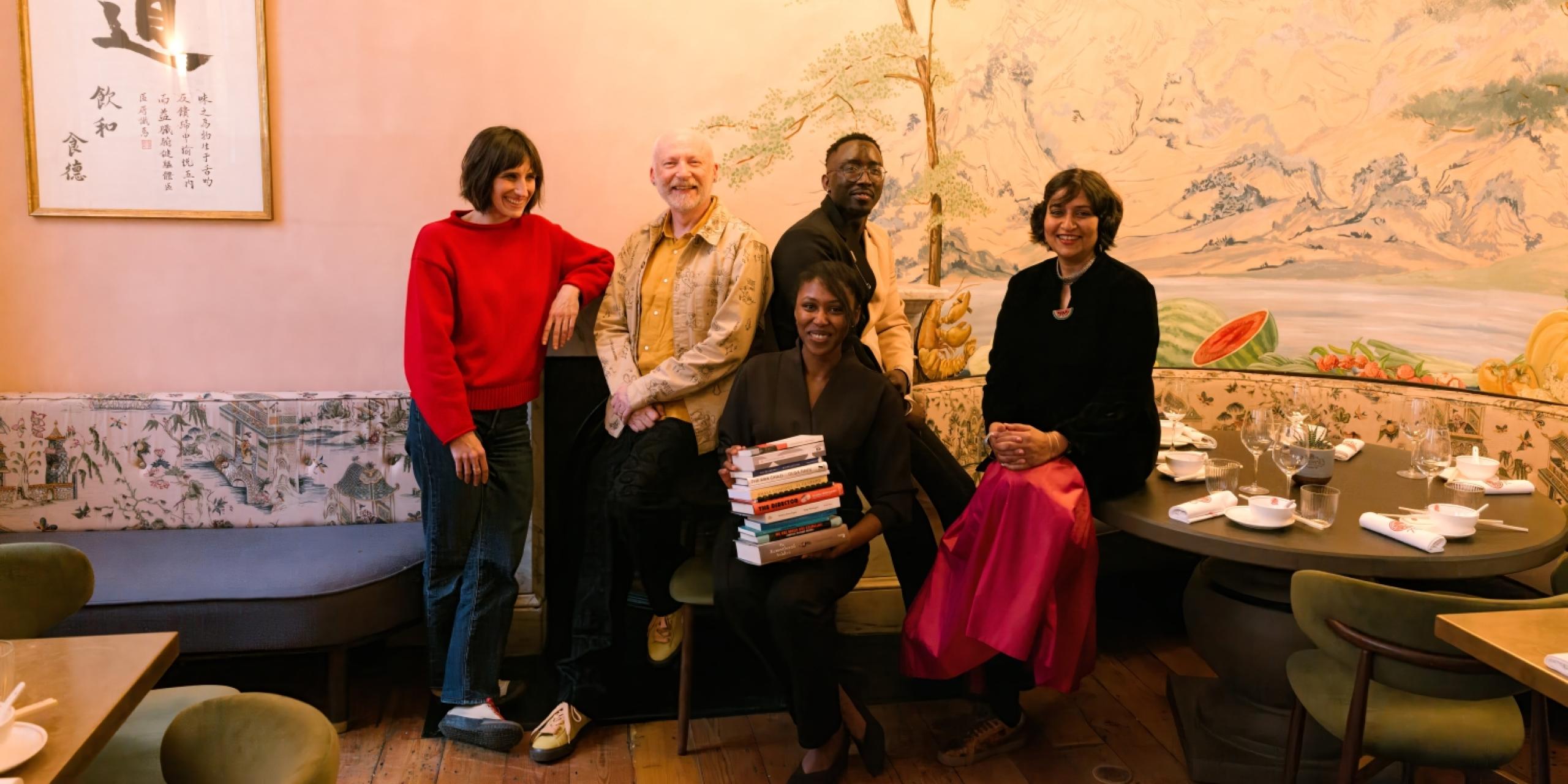 A group portrait of International Booker Prize 2026 judges Natasha Brown, Marcus du Sautoy, Sophie Hughes, Troy Onyango and Nilanjana S. Roy. holding the longlisted books