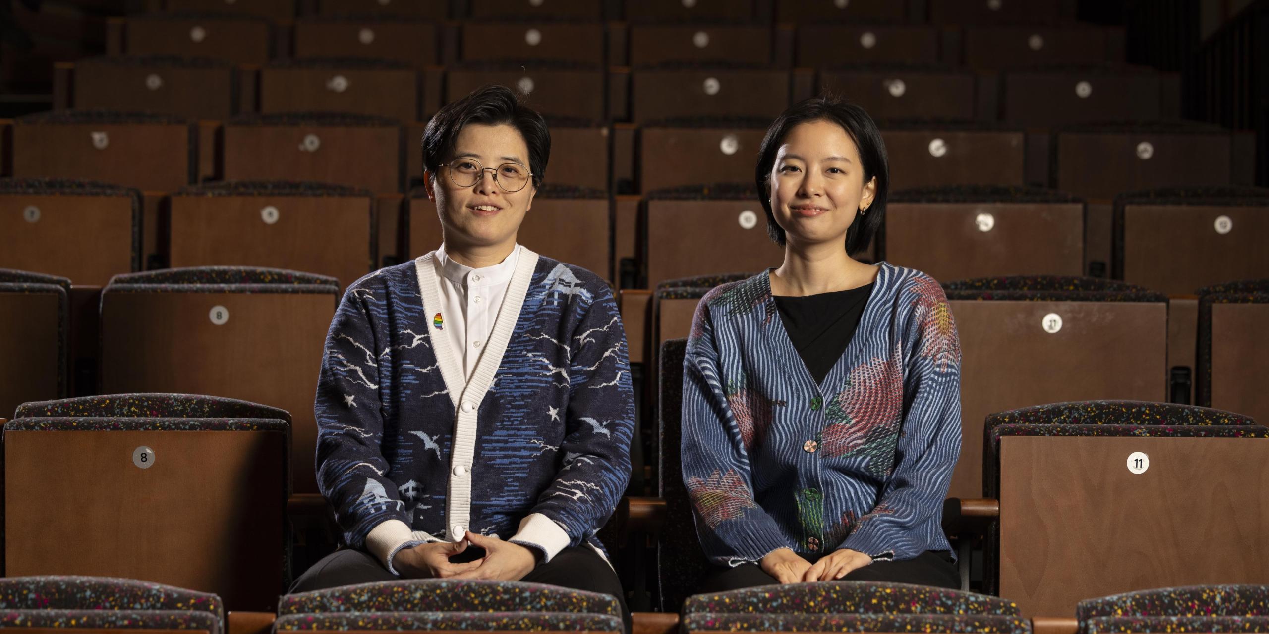 Two individuals sit in an auditorium. One has short hair and glasses. One has longer hair. Both are wearing cardigans and smiling at the camera. 
