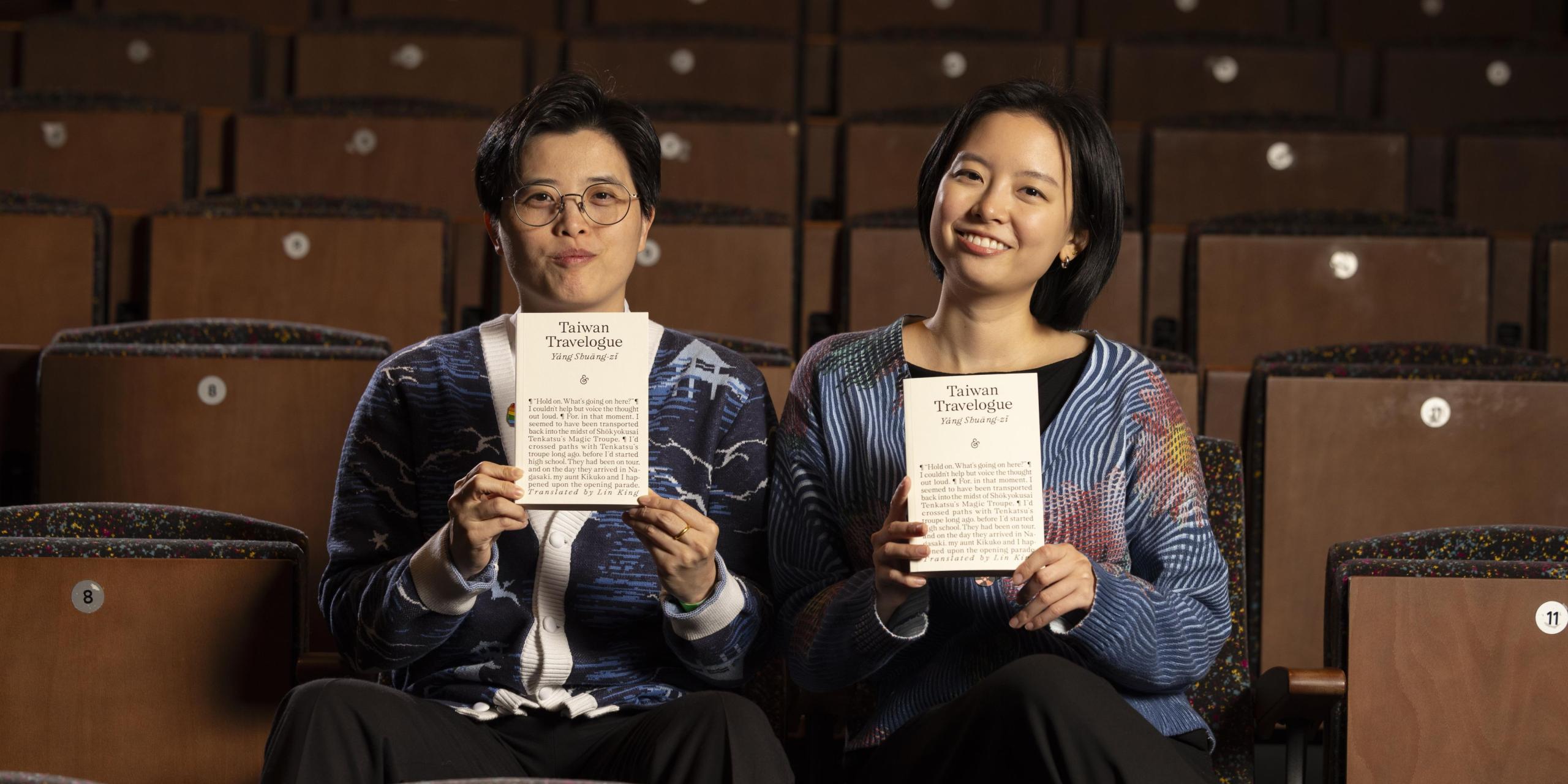 Two individuals sit in an auditorium holding a copy of Taiwan Travelogue.One has short hair and glasses. One has longer hair. Both are wearing cardigans and smiling at the camera. 