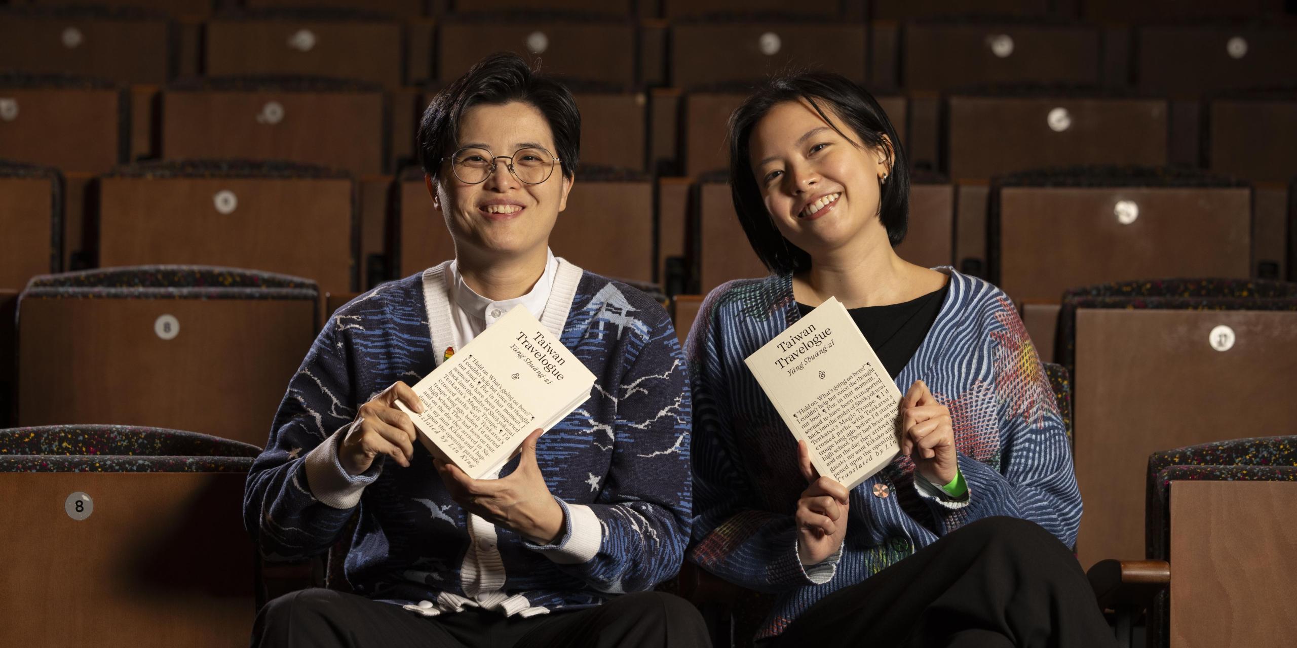 Two individuals sit in an auditorium holding up a copy of Taiwan Travelogue. One has short hair and glasses. One has longer hair. Both are wearing cardigans and smiling at the camera. 